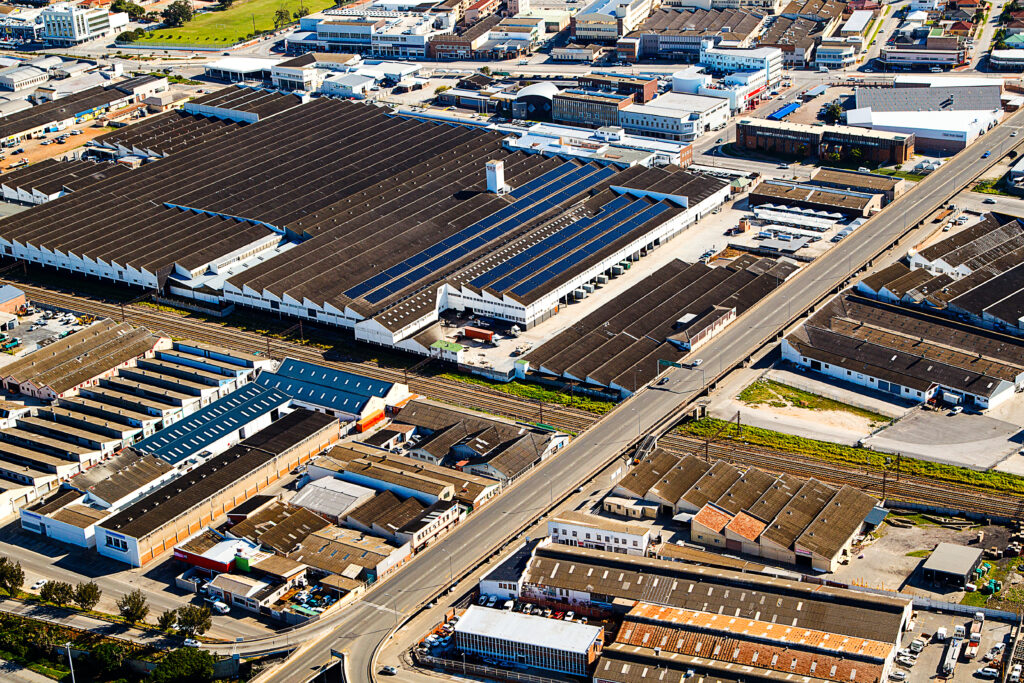 An aerial shot captures an industrial area filled with various factory buildings, some featuring solar panels on their roofs. A long road cuts through the center, paralleled by railroad tracks, under a clear blue sky.