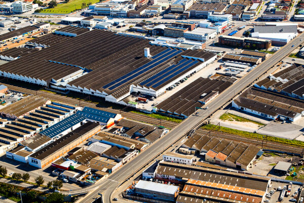 An aerial shot captures an industrial area filled with various factory buildings, some featuring solar panels on their roofs. A long road cuts through the center, paralleled by railroad tracks, under a clear blue sky.